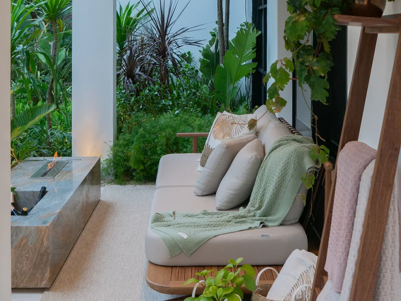 Queimador QS-2000 QS-2000-A da Somer com chama acesa embutido em mesa de pedra projetada em varanda residencial de alto padrão, com sofá em primeiro plano e vegetação tropical ao fundo.