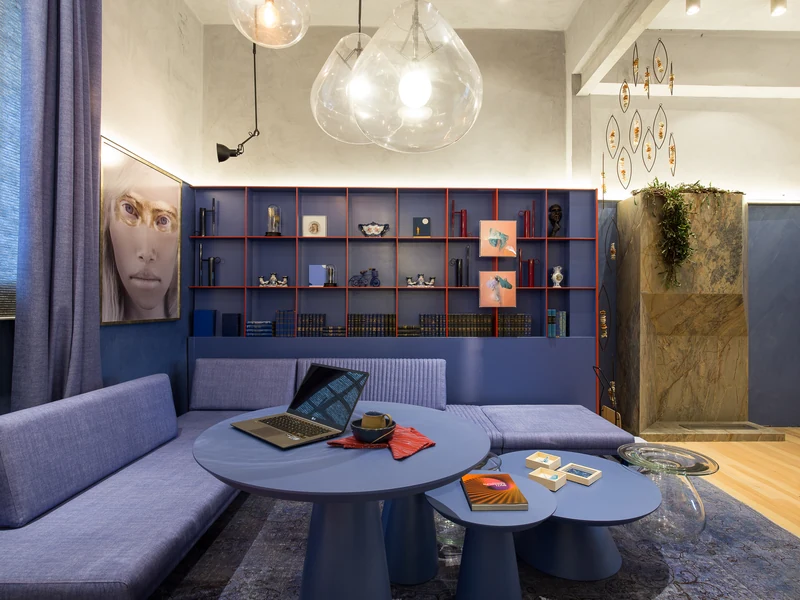 Queimador QS-3000 QS-3000-A da Somer com chama acesa embutido em painel de pedra em sala-lounge de exposição, com sofá em L azul-violeta, prateleiras com objetos, abajures pendentes em vidro soprado.