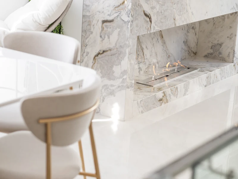 Detalhe do queimador QS-3500 QS-3500-A da Somer com chama acesa em duas seções embutido em painel de mármore Calacatta em sala de estar de alto padrão com cadeiras brancas com detalhes dourados.