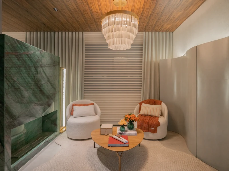 Queimador QS-3500 QS-3500-A da Somer embutido em painel de mármore verde em sala intimista de leitura com duas poltronas brancas, mesa de centro em madeira, lustre de cristal e teto ripado.