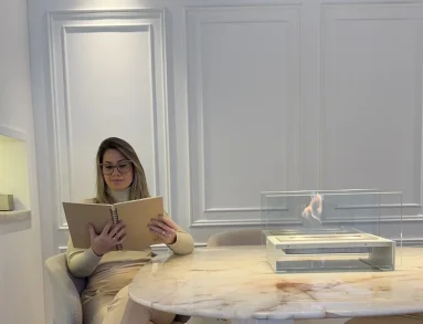 Mulher lendo ao lado da Lareira Ecológica GO! Branca da Somer com chama viva sobre mesa de mármore em showroom branco